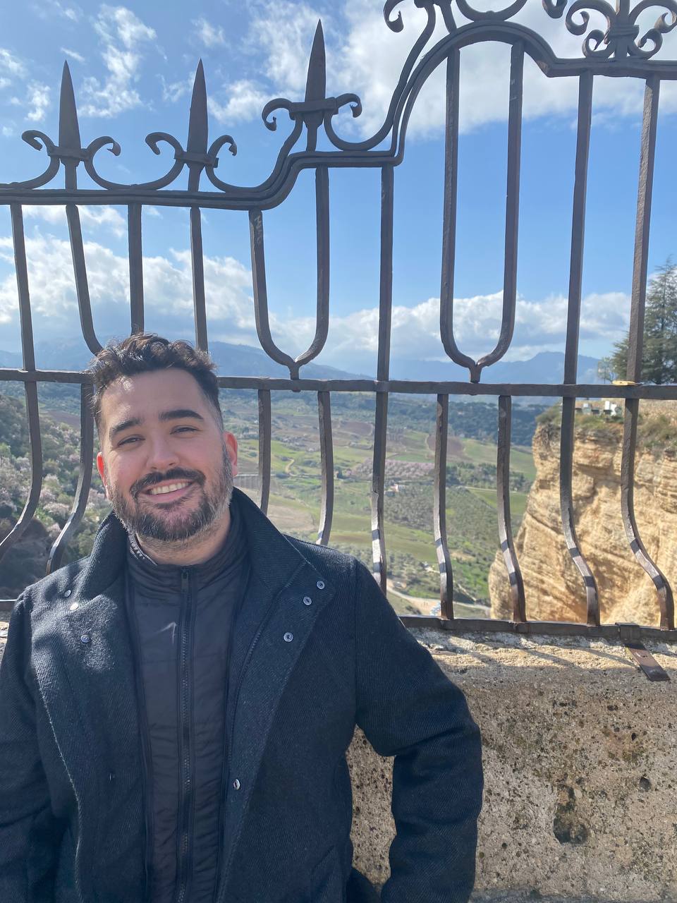 Alex smiling in front of an iron railing, with hills and farmland behind